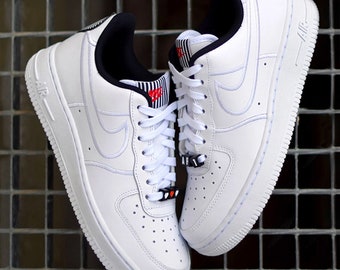 May include: White leather low-top sneakers with black and red accents. The sneakers have a white swoosh logo and the word "AIR" on the back.