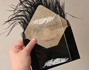 May include: A black envelope with black ostrich feathers, containing a gold glitter card with a white feather illustration and the text "gift card" and "SWANTOR knitted clothes" printed in black, along with a clear acrylic overlay.