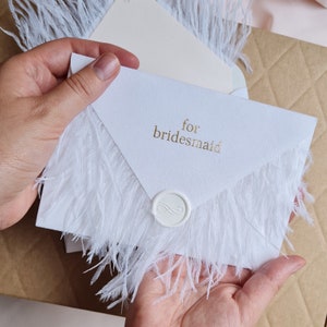 May include: White envelope with a gold inscription that reads "for bridesmaid" and a white wax seal. The envelope is decorated with white feathers.