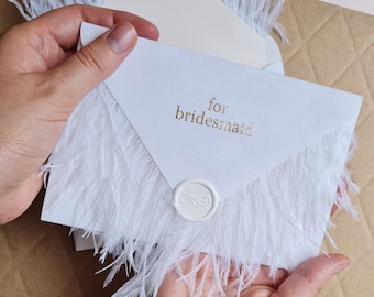 May include: White envelope with a gold inscription that reads "for bridesmaid" and a white wax seal. The envelope is decorated with white feathers.