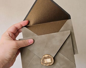 May include: A sage green envelope with a pointed flap, featuring a metallic bronze inner lining. A smaller sage green envelope is layered on top, secured with a rectangular gold wax seal that displays an intricate, ornate frame design.