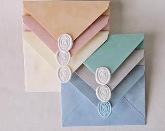May include: Two stacks of envelopes in pastel shades of peach, pink, cream, green, gray, and blue. Each stack is sealed with three vertical white wax seals featuring an embossed cameo profile design on the pointed envelope flaps.