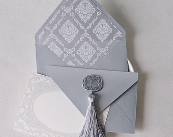 May include: A light gray envelope with a pointed flap featuring a white damask pattern on the interior lining. The envelope is sealed with a silver wax seal and a matching silver tassel. A rectangular card with a decorative border is visible.