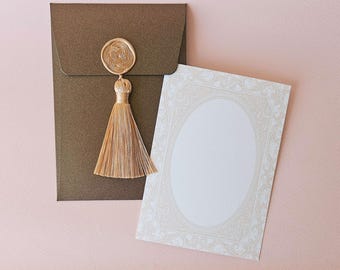 May include: A dark bronze envelope featuring a circular gold wax seal with a woven knot design and a long, light gold tassel attached. Beside the envelope is a rectangular white card with a light gold ornate border and a central oval frame.