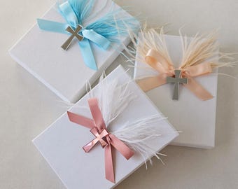 May include: Three rectangular white gift boxes, each decorated with a satin ribbon bow, a cluster of soft feathers, and a metallic cross charm. The ribbons and feathers are in light blue, peach, and rose gold colors, centered on each box lid.
