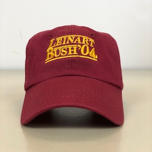 May include: A maroon baseball cap with a gold embroidered logo that reads "LEINART BUSH '04".