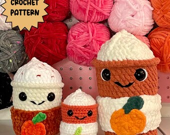 May include: Three crocheted amigurumi pumpkin spice latte cup characters. The largest cup is brown with a white lid and a white crocheted pumpkin. The middle cup is white with a brown crocheted pumpkin and a brown lid. The smallest cup is white with a red crocheted pumpkin and a red lid.