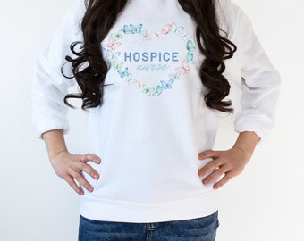 May include: White sweatshirt featuring the text "HOSPICE nurse" within a heart-shaped arrangement of pastel butterflies. The long-sleeved top is made of a soft material and is worn with blue jeans.