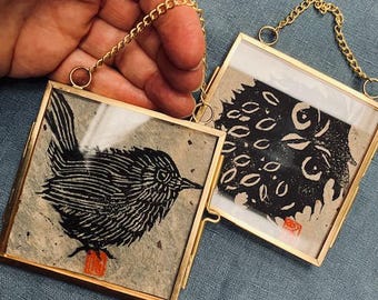 May include: Two square, gold-framed art pieces with black and white bird and abstract designs. The art pieces are held by a gold chain. The bird art has a red square at the bottom. The background is a light gray.