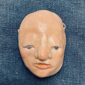 May include: A hand-crafted clay mask depicting a human face with a large nose and closed eyes. The mask is painted in shades of peach and brown.