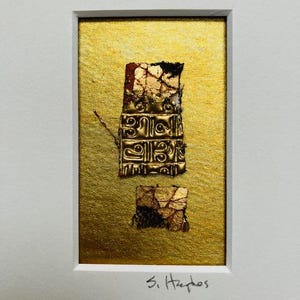 May include: A gold-toned, textured, rectangular piece of art with a red and black design. The art is framed and has the text "3. Hephaestos" written below it.