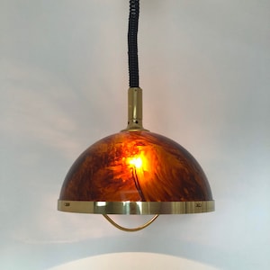 May include: A vintage pendant light with an amber-coloured, domed shade. The shade has a marbled effect and is trimmed with a brass band. A black, coiled cord suspends the light from the ceiling, with a brass-coloured stem.