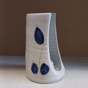 May include: A ceramic napkin holder with a light gray finish. The holder features a cut-out design with three blue leaf-shaped accents. The holder has a curved opening on one side and a flat base.
