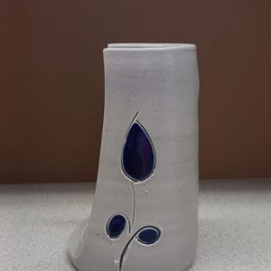 May include: A handmade, off-white ceramic vase with a dark blue floral design. The vase has a unique, curved shape and a flat base. The floral design includes a large teardrop shape and two smaller circles. The vase is suitable for home decor.