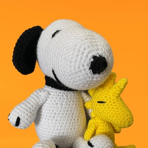 May include: White crocheted Snoopy plush toy with black ears and nose, sitting with a yellow crocheted Woodstock plush toy.