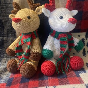 May include: Two crocheted reindeer toys, one brown and one white, both wearing green and red striped scarves. The reindeer are sitting on a red and white plaid blanket.