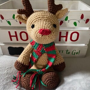 May include: A crocheted reindeer toy wearing a green and red striped scarf with tassels. The reindeer is sitting in front of a white wooden box with the word "HO HO" written on it.