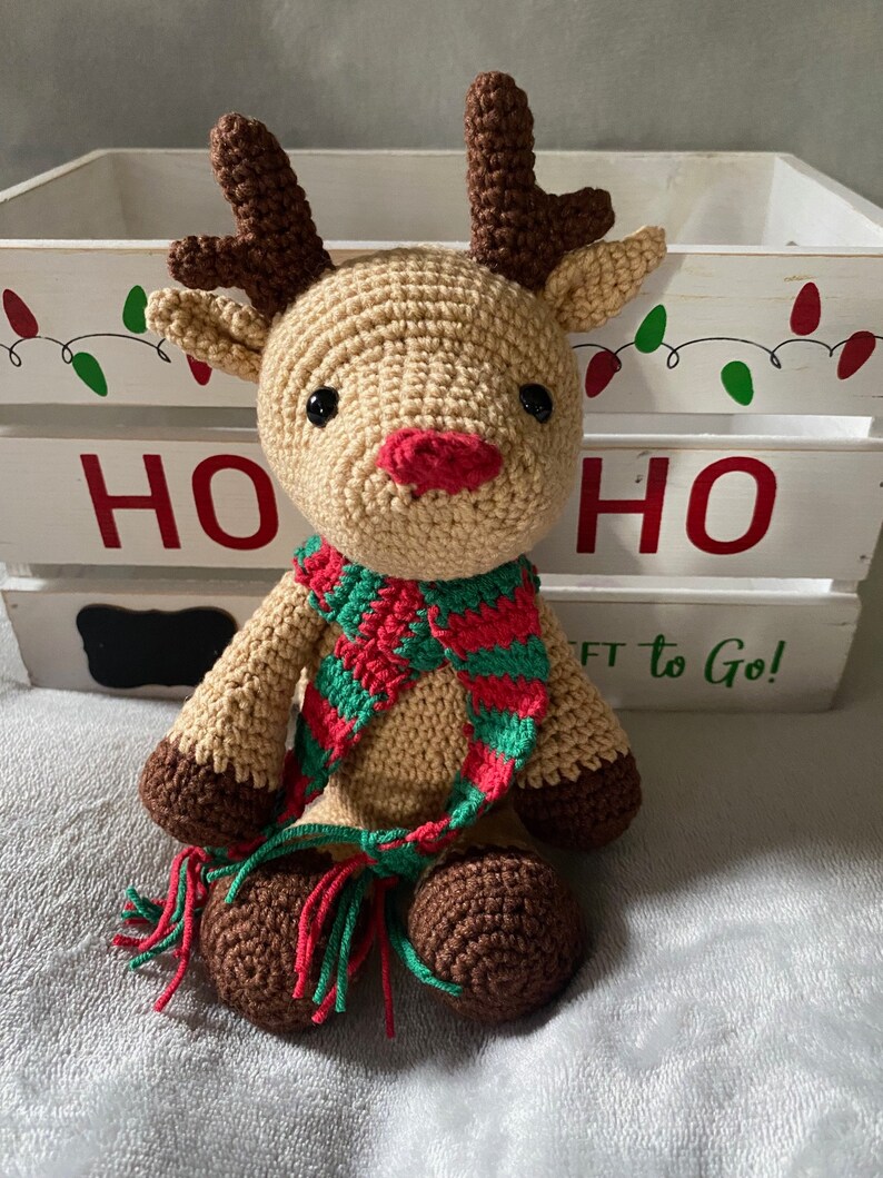 May include: A crocheted reindeer toy wearing a green and red striped scarf with tassels. The reindeer is sitting in front of a white wooden box with the word "HO HO" written on it.