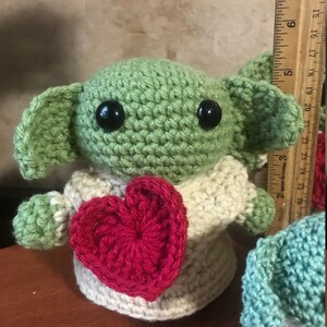 Puede incluir: Un juguete de peluche verde tejido a crochet con grandes ojos negros, sosteniendo un coraz&oacute;n rojo tejido a crochet. El juguete lleva un su&eacute;ter tejido a crochet de color crema.