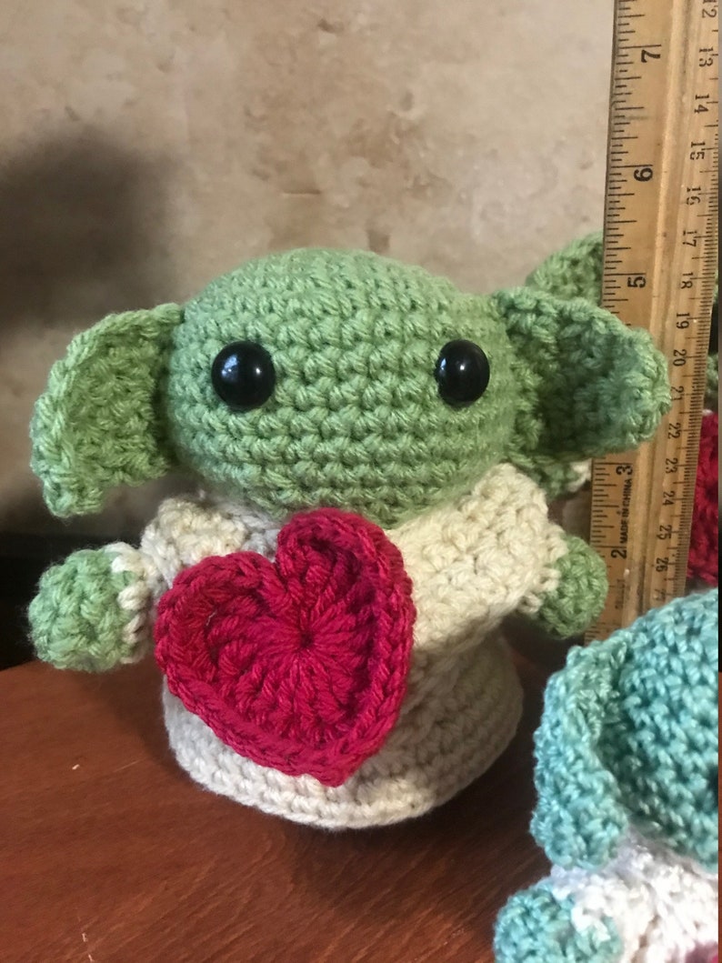 Puede incluir: Un juguete de peluche verde tejido a crochet con grandes ojos negros, sosteniendo un coraz&oacute;n rojo tejido a crochet. El juguete lleva un su&eacute;ter tejido a crochet de color crema.