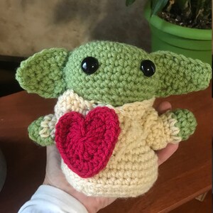 Puede incluir: Un juguete de peluche verde tejido a crochet con grandes ojos negros y un coraz&oacute;n rojo en el pecho. El juguete lleva un su&eacute;ter tejido a crochet de color crema.