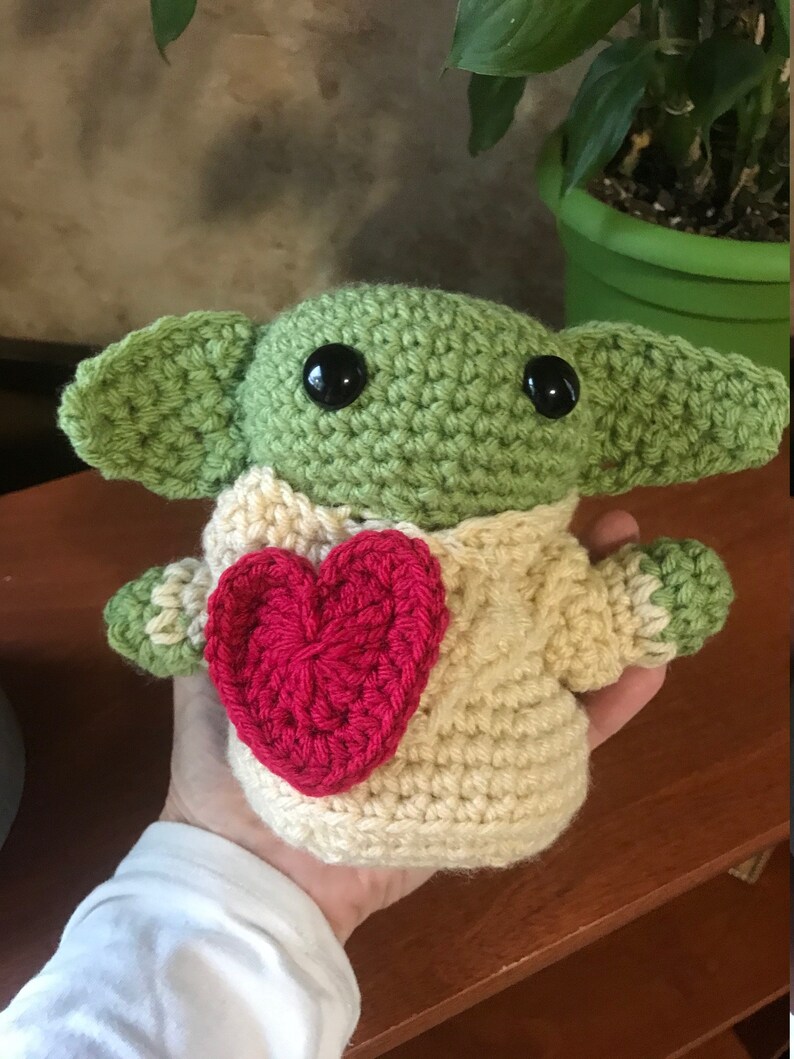 Puede incluir: Un juguete de peluche verde tejido a crochet con grandes ojos negros y un coraz&oacute;n rojo en el pecho. El juguete lleva un su&eacute;ter tejido a crochet de color crema.