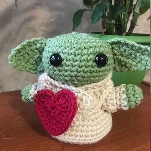 Puede incluir: Un juguete de peluche verde tejido a crochet con grandes ojos negros y un coraz&oacute;n rojo en sus manos. El juguete lleva un su&eacute;ter tejido a crochet de color crema.