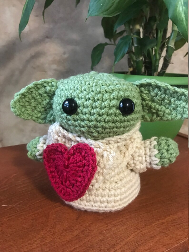 Puede incluir: Un juguete de peluche verde tejido a crochet con grandes ojos negros y un coraz&oacute;n rojo en sus manos. El juguete lleva un su&eacute;ter tejido a crochet de color crema.