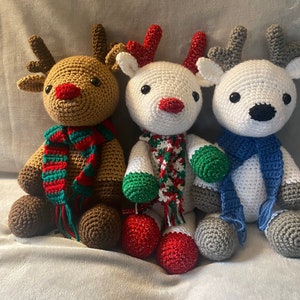 May include: Three crocheted reindeer dolls. One is brown with a red nose and a green and red scarf. The second is white with a red nose and a green and red scarf. The third is white with a black nose and a blue scarf.
