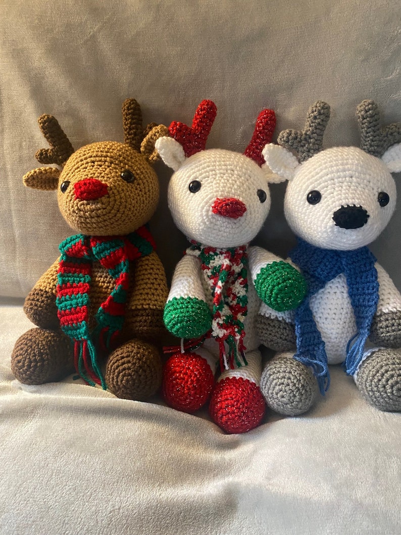 May include: Three crocheted reindeer dolls. One is brown with a red nose and a green and red scarf. The second is white with a red nose and a green and red scarf. The third is white with a black nose and a blue scarf.