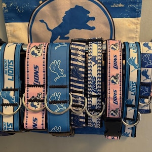 May include: A set of seven dog collars featuring a Detroit Lions theme. The collars are made of blue, pink, and white ribbon with a variety of designs including the team logo, the words "Detroit Lions", and a lion head.