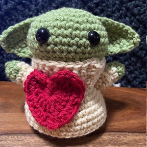Puede incluir: Un juguete de peluche verde y beige tejido a crochet con grandes ojos negros, que lleva un su&eacute;ter beige con un coraz&oacute;n rojo en la parte delantera.