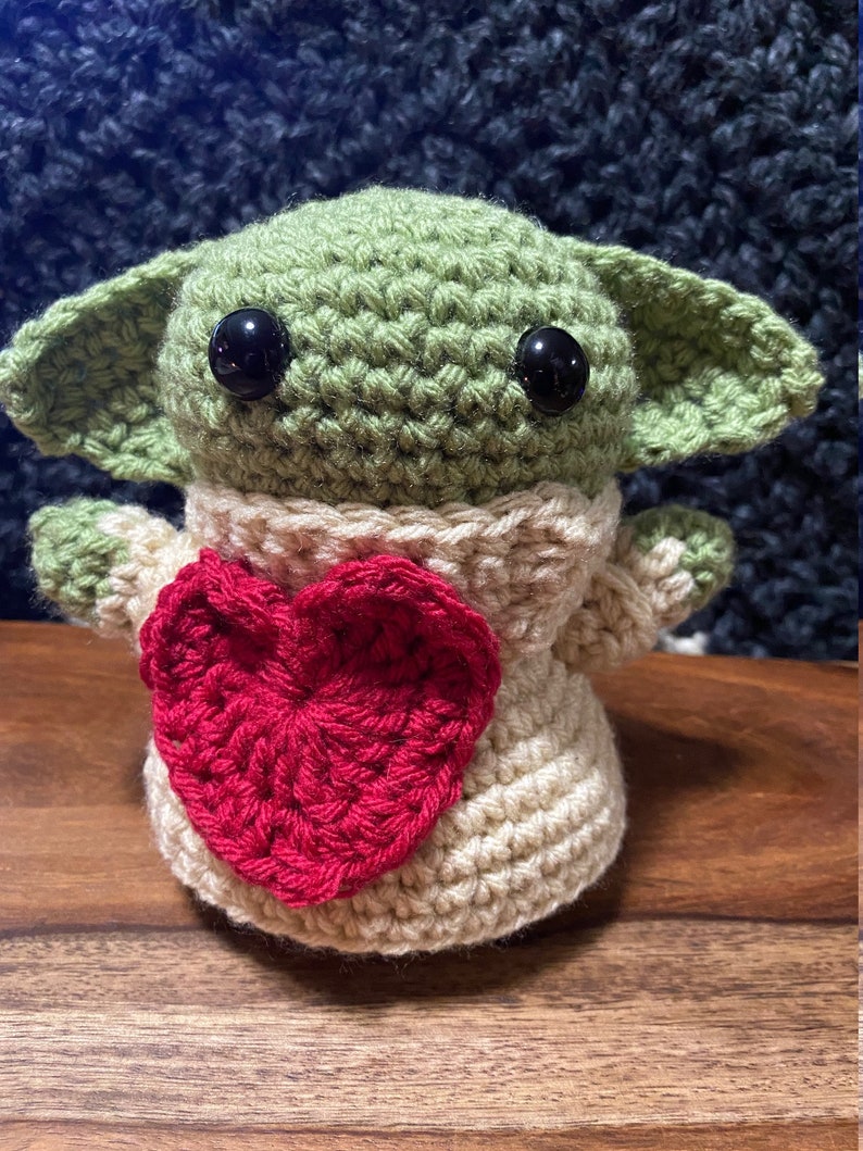 Puede incluir: Un juguete de peluche verde y beige tejido a crochet con grandes ojos negros, que lleva un su&eacute;ter beige con un coraz&oacute;n rojo en la parte delantera.