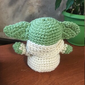 Puede incluir: Un mu&ntilde;eco de beb&eacute; Yoda de crochet de color verde y crema con orejas grandes. El mu&ntilde;eco est&aacute; sentado sobre una superficie de madera.