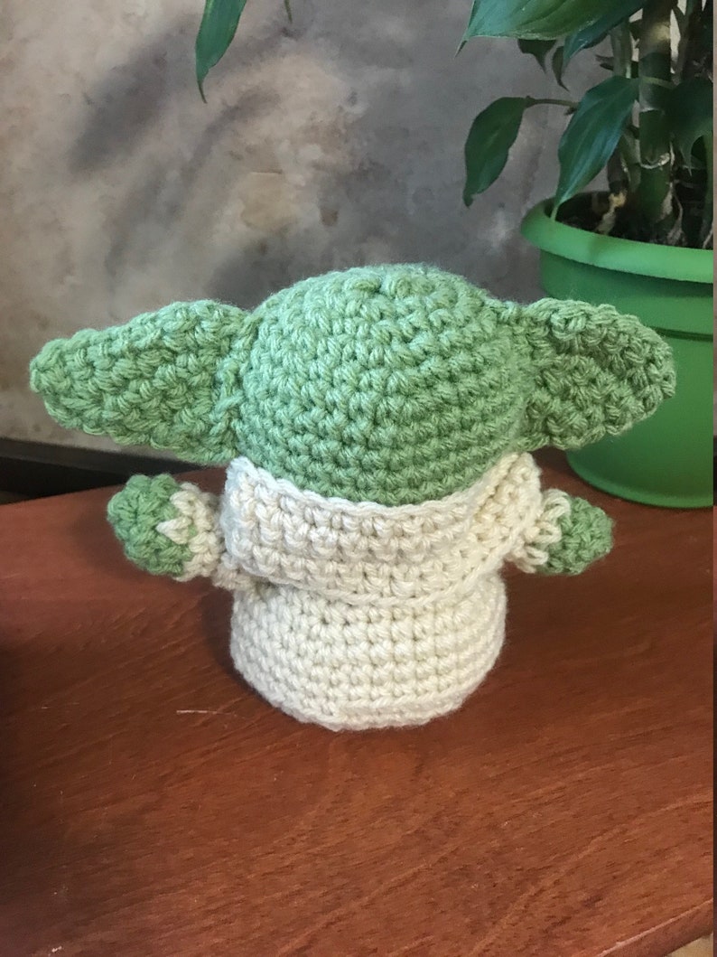 Puede incluir: Un mu&ntilde;eco de beb&eacute; Yoda de crochet de color verde y crema con orejas grandes. El mu&ntilde;eco est&aacute; sentado sobre una superficie de madera.