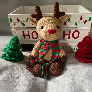 May include: A crocheted reindeer toy with a red and green striped scarf, sitting in front of a white wooden crate with the word "HO HO" painted on it. There are two small crocheted Christmas trees on either side of the reindeer.