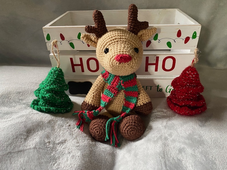 May include: A crocheted reindeer toy with a red and green striped scarf, sitting in front of a white wooden crate with the word "HO HO" painted on it. There are two small crocheted Christmas trees on either side of the reindeer.