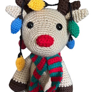 May include: A handmade crochet reindeer with a red nose, brown antlers, and a striped red and green scarf. The reindeer is adorned with colorful crochet Christmas lights, including yellow, blue, and green.