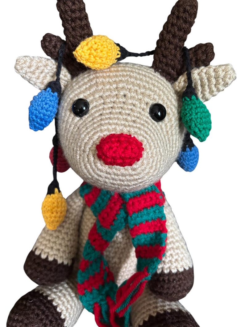 May include: A handmade crochet reindeer with a red nose, brown antlers, and a striped red and green scarf. The reindeer is adorned with colorful crochet Christmas lights, including yellow, blue, and green.