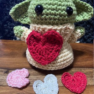 Puede incluir: Un juguete de peluche verde tejido a crochet con grandes ojos negros, que lleva un su&eacute;ter tejido a crochet de color crema con un coraz&oacute;n tejido a crochet rojo en la parte delantera. Tres corazones tejidos a crochet en rosa, blanco y rojo est&aacute;n sobre una superficie de madera.