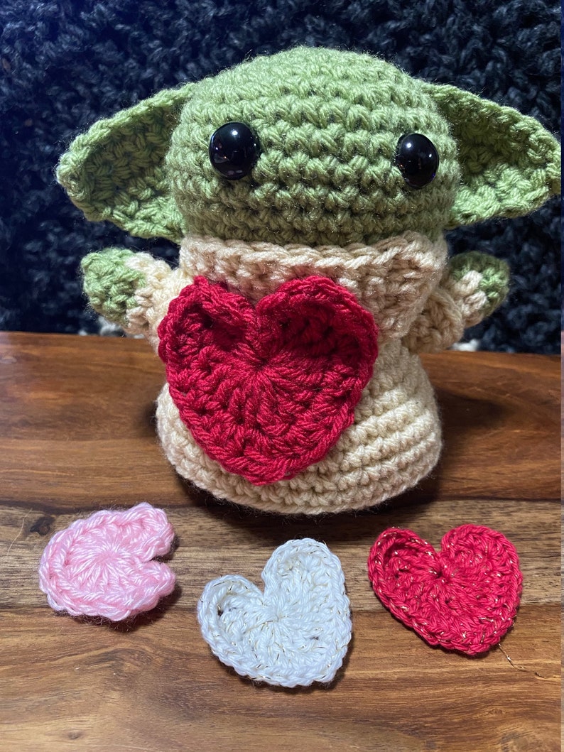 Puede incluir: Un juguete de peluche verde tejido a crochet con grandes ojos negros, que lleva un su&eacute;ter tejido a crochet de color crema con un coraz&oacute;n tejido a crochet rojo en la parte delantera. Tres corazones tejidos a crochet en rosa, blanco y rojo est&aacute;n sobre una superficie de madera.