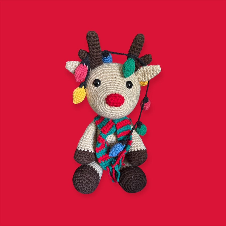 May include: A crocheted reindeer with brown antlers, a red nose, and a colorful scarf. The reindeer is adorned with a string of decorative lights in various colors. The background is a solid red.
