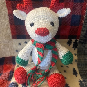 May include: White crocheted reindeer with red antlers and a green and red striped scarf. The reindeer is sitting on a white and red plaid blanket.
