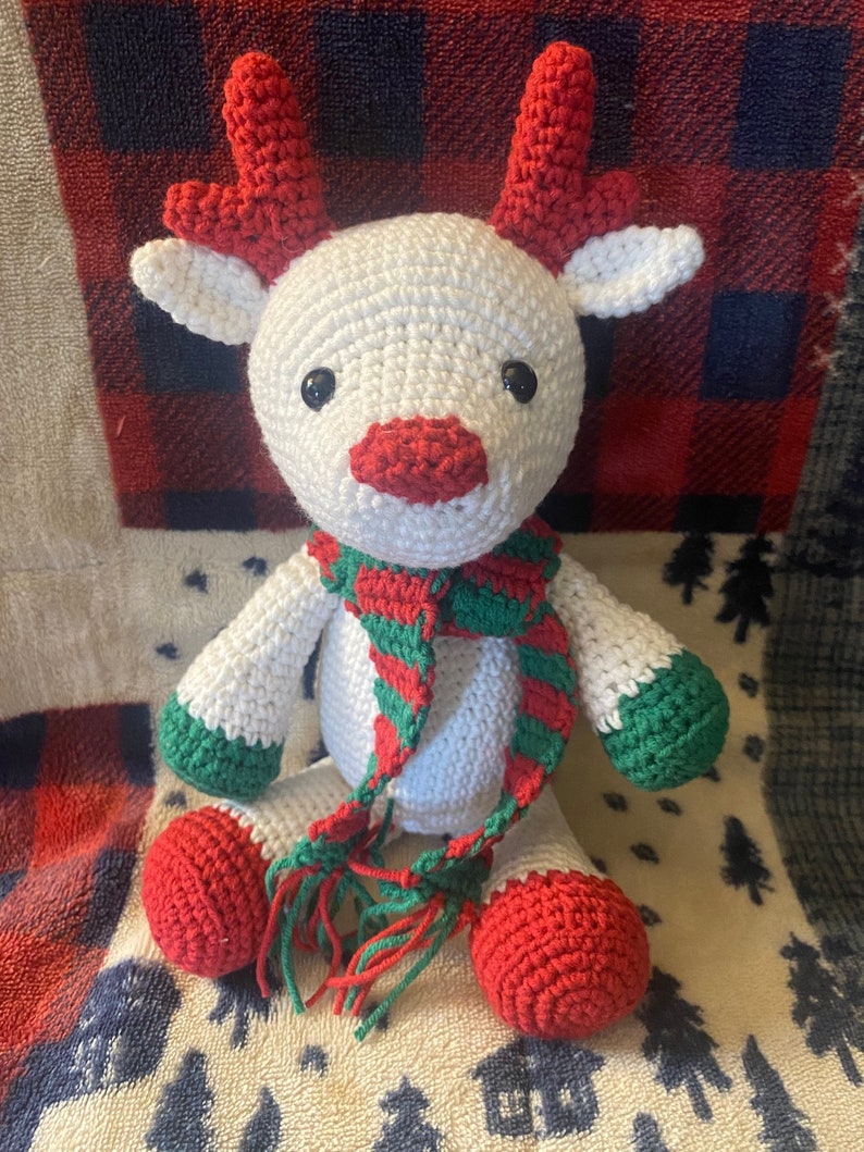 May include: White crocheted reindeer with red antlers and a green and red striped scarf. The reindeer is sitting on a white and red plaid blanket.