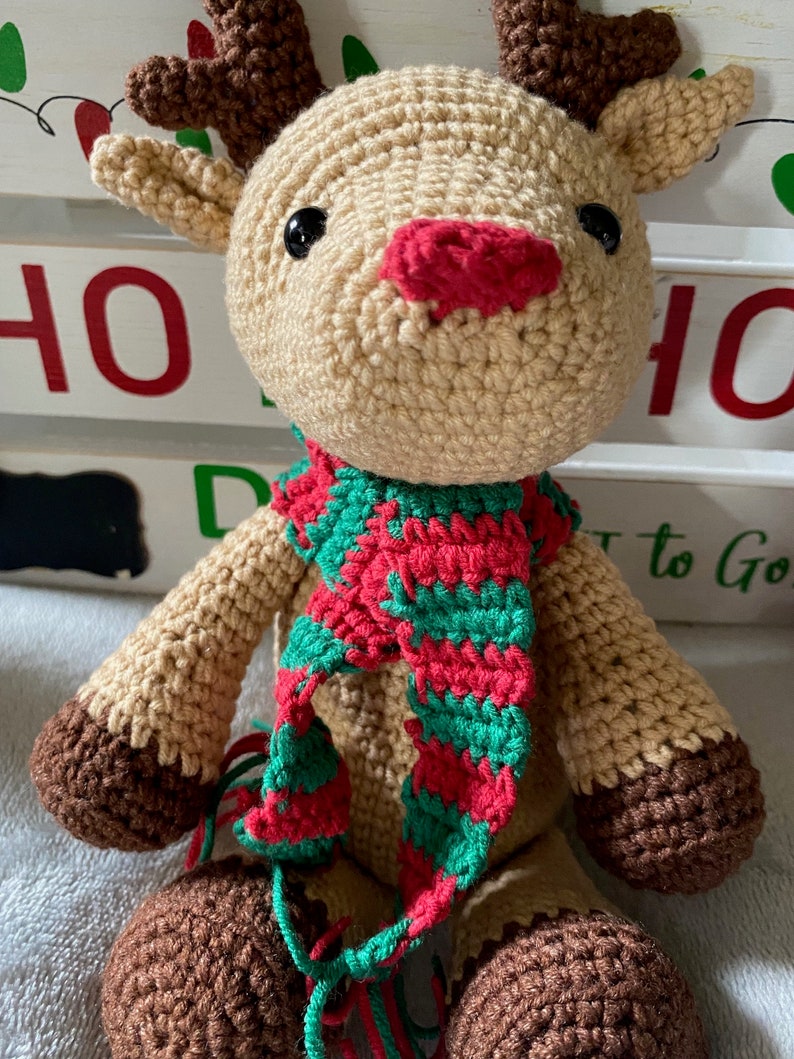 May include: A crocheted reindeer toy with brown antlers, a red nose, and a green and red striped scarf.