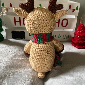 May include: A crocheted reindeer toy with brown antlers, a tan body, and a red and green striped scarf. The reindeer is sitting on a white fuzzy surface in front of a white wooden crate with the word "HO" written on it.