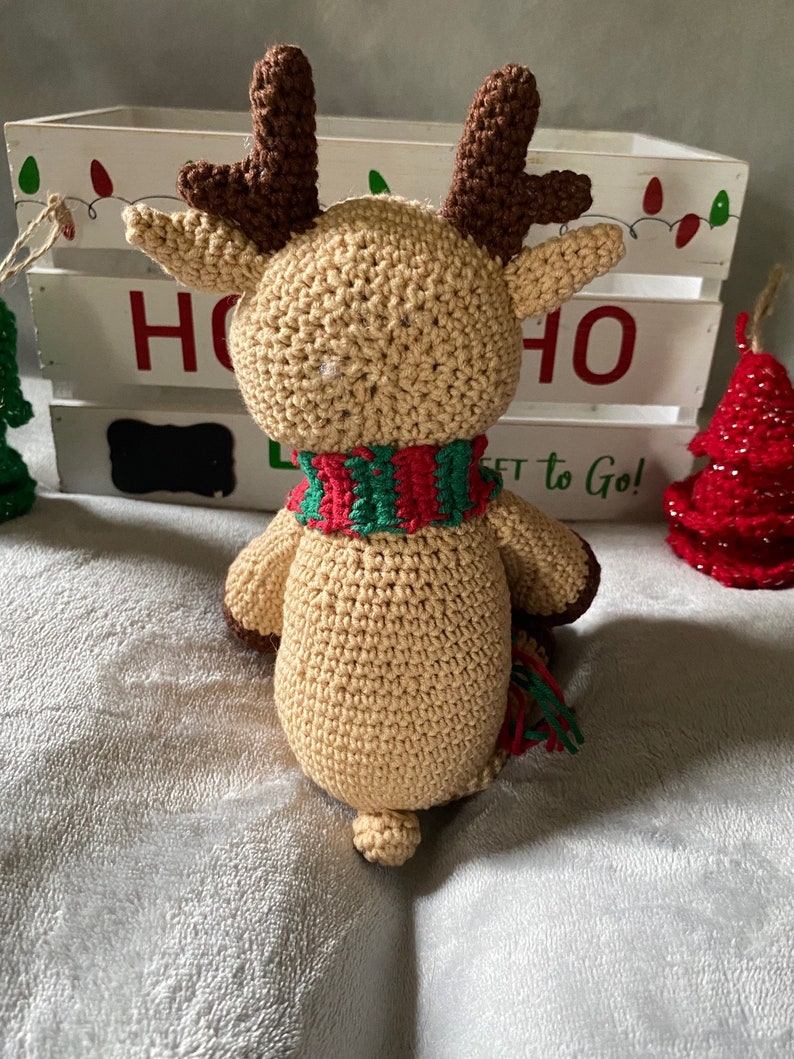 May include: A crocheted reindeer toy with brown antlers, a tan body, and a red and green striped scarf. The reindeer is sitting on a white fuzzy surface in front of a white wooden crate with the word "HO" written on it.