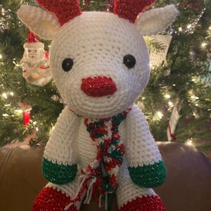 May include: A handmade white crochet reindeer with red antlers, nose, and feet, and green accents on the legs. It has a festive red, green, and white scarf. The reindeer is a Christmas decoration.