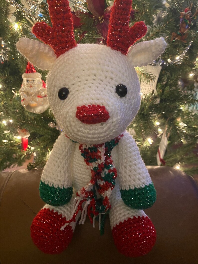May include: A handmade white crochet reindeer with red antlers, nose, and feet, and green accents on the legs. It has a festive red, green, and white scarf. The reindeer is a Christmas decoration.