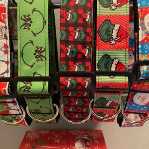 May include: A collection of festive dog collars with various holiday-themed patterns. Designs include gnomes, the Grinch, plaid with gifts, Santa Claus, and snowmen. Each collar has a black buckle and a silver D-ring for leash attachment.
